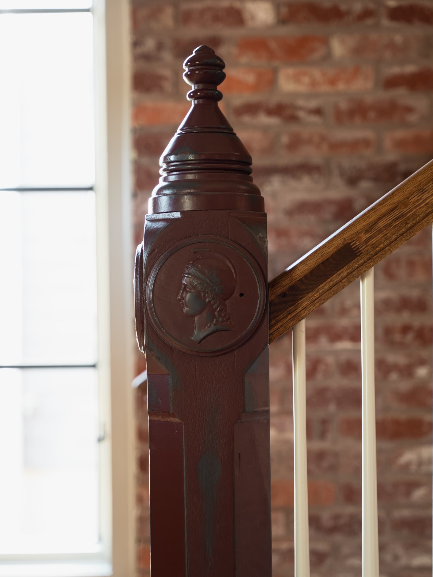 A handcrafted newel post with a classical female-profile medallion at the foot of an interior stair, brick wall behind